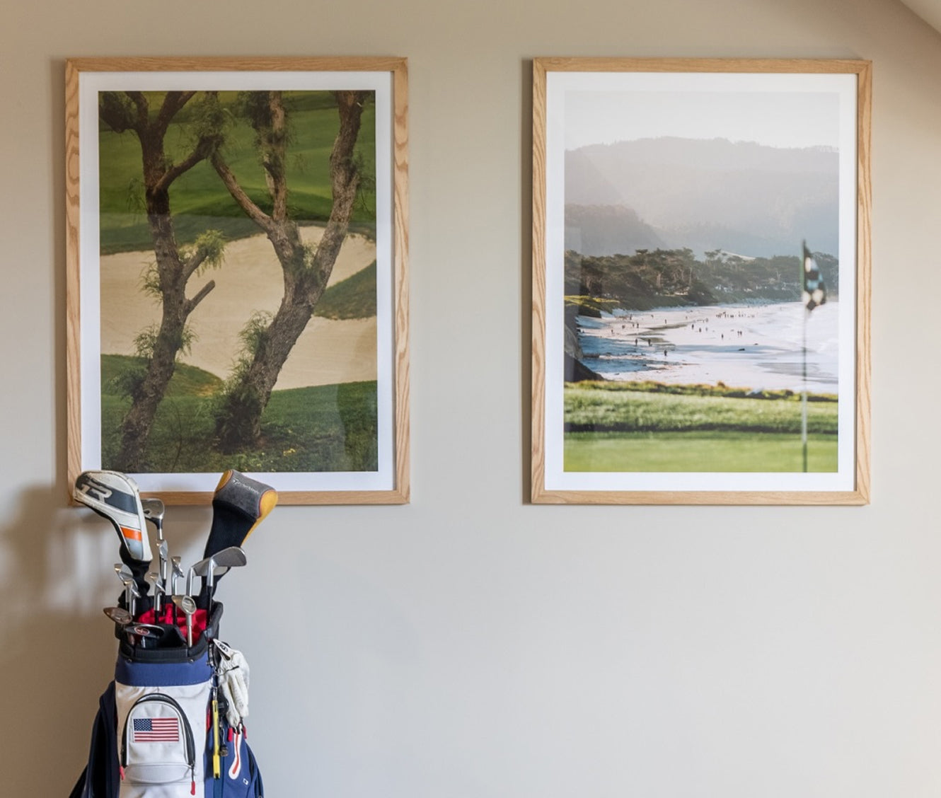 Two framed golf course art prints hanging on a wall above a golf bag